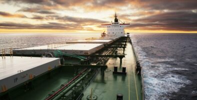 A green bulk carrier sails forward on calm waters with a mild yellow sunset behind grey clouds