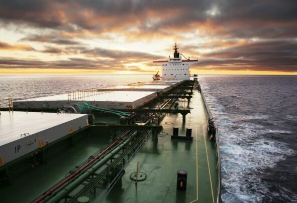 A green bulk carrier sails forward on calm waters with a mild yellow sunset behind grey clouds