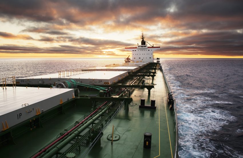 A green bulk carrier sails forward on calm waters with a mild yellow sunset behind grey clouds