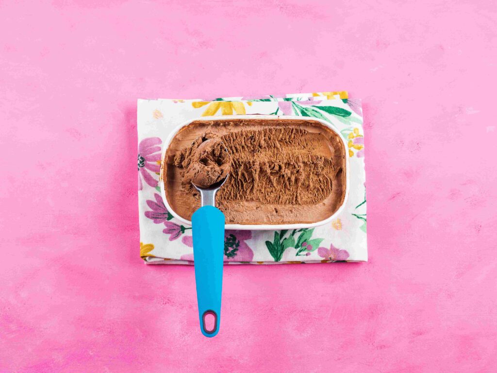 Ice cream scooper in a tub of chocolate ice cream that is atop a floral napkin on a pink table.